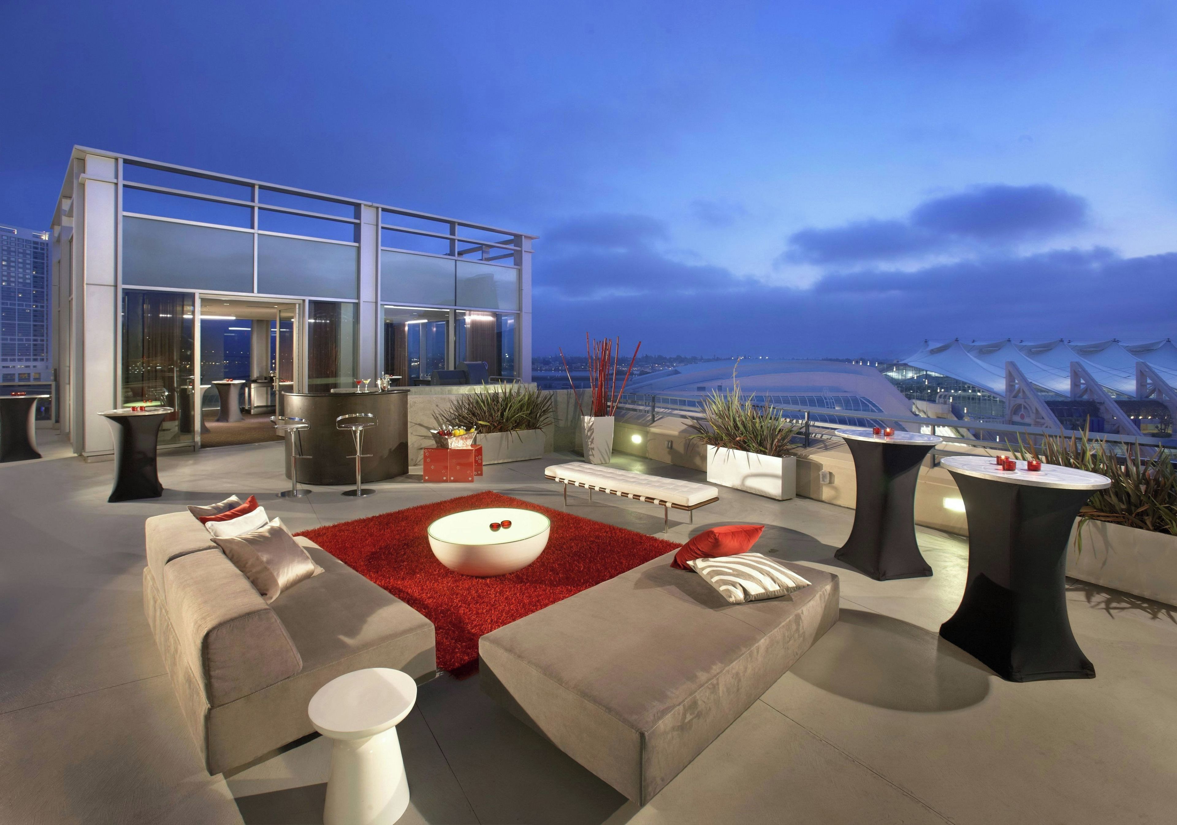 A rooftop lounge with modern furniture, a glass entrance, and a view of a bridge and cityscape.