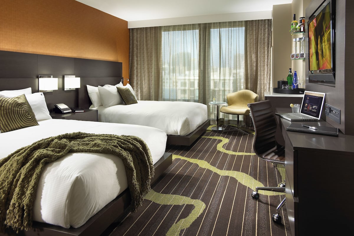 Habitación de hotel con dos camas, un escritorio con una computadora portátil, una silla y vistas a la ciudad a través de amplias ventanas.