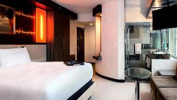 Ein modernes Hotelzimmer mit einem großen Bett, einem kleinen Tisch, einem Fernseher und einem durch eine Glastür sichtbaren Badezimmer.