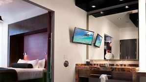 Quarto de hotel moderno com uma cama, uma banheira grande, uma toucador com espelho e duas televisões fixadas na parede exibindo uma cena de praia.