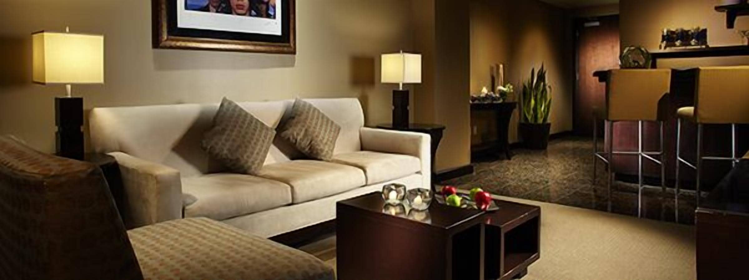 A hotel room with a sofa, two lamps, a coffee table with fruits, and a bar area in the background.