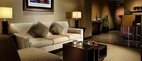 A hotel room with a sofa, two lamps, a coffee table with fruits, and a bar area in the background.