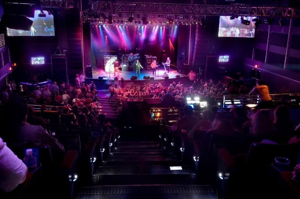 Un recinto para conciertos con asientos escalonados, un escenario con artistas y pantallas grandes mostrando el evento.