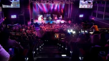 Un recinto para conciertos con asientos escalonados, un escenario con artistas y pantallas grandes mostrando el evento.