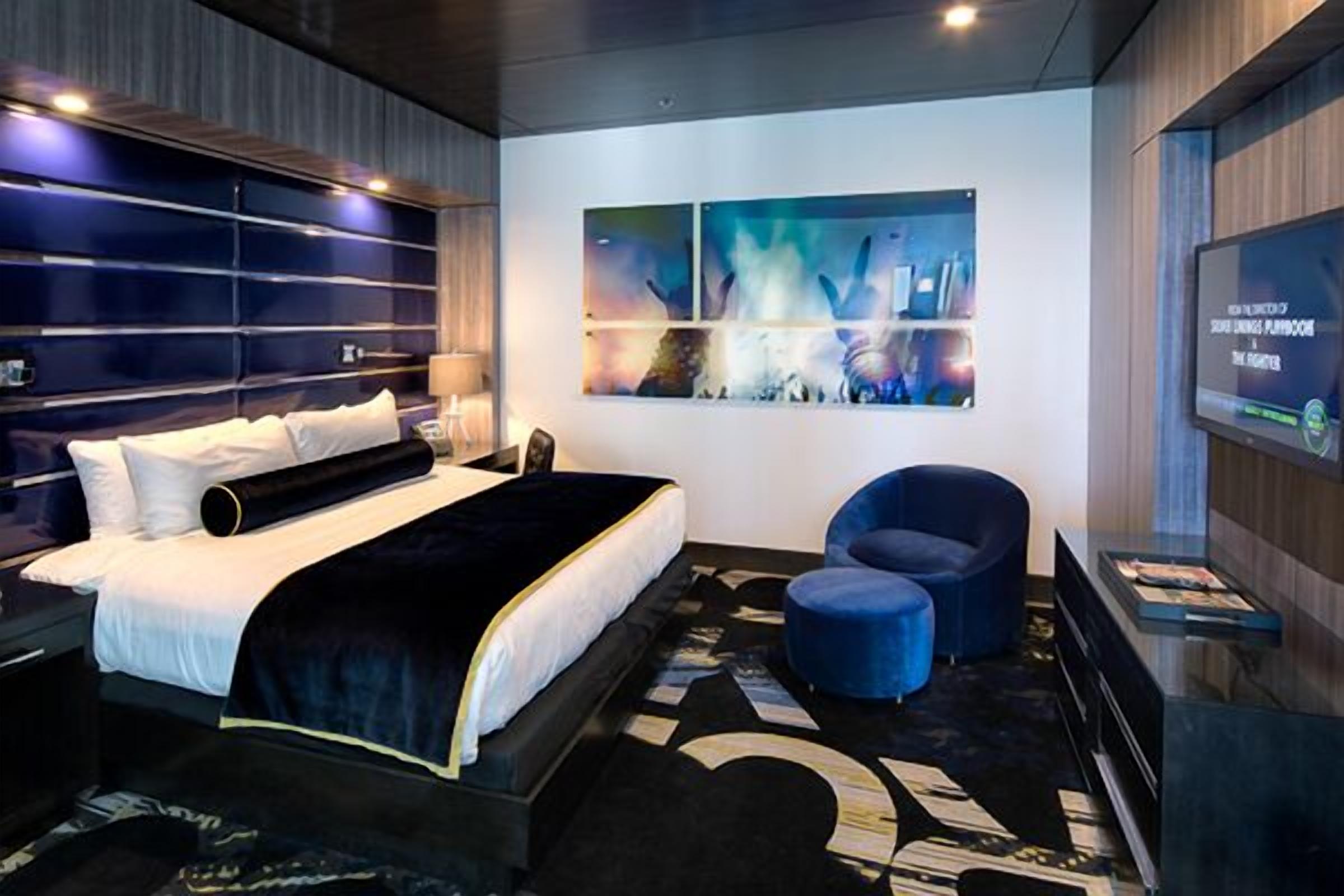 A modern hotel room with a large bed, a blue armchair, a small ottoman, and a wall-mounted TV.