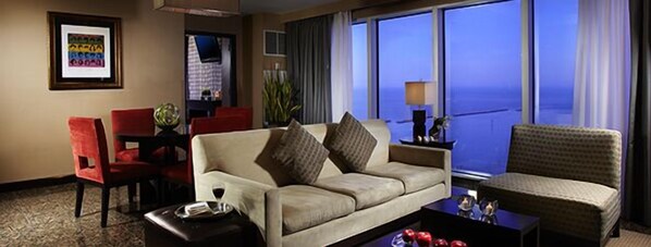 Habitación de hotel con sofá, dos sillones, una mesa de centro y vistas al océano a través de amplias ventanas.