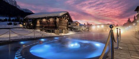 A wooden cabin with a hot tub in the foreground, surrounded by snow and mountains.