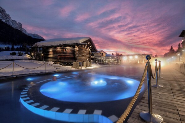 Una cabaña de madera con jacuzzi en primer plano, rodeada de nieve y montañas.