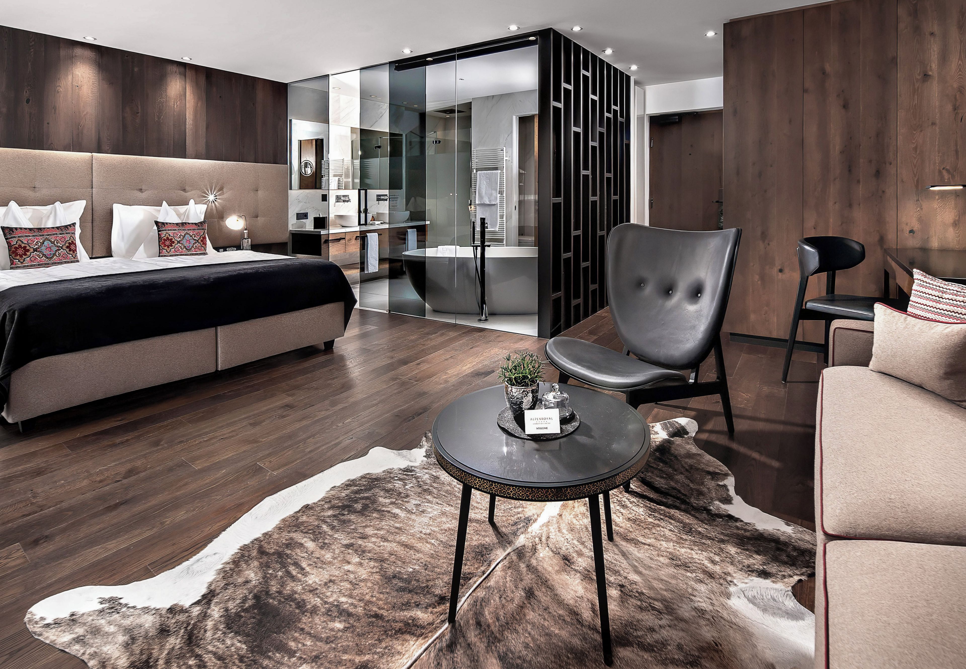 Ein modernes Schlafzimmer mit einem großen Bett, einer Sitzecke inklusive Sessel, einem kleinen Tisch und einem ein Glasfenster zeigenden Badezimmer.