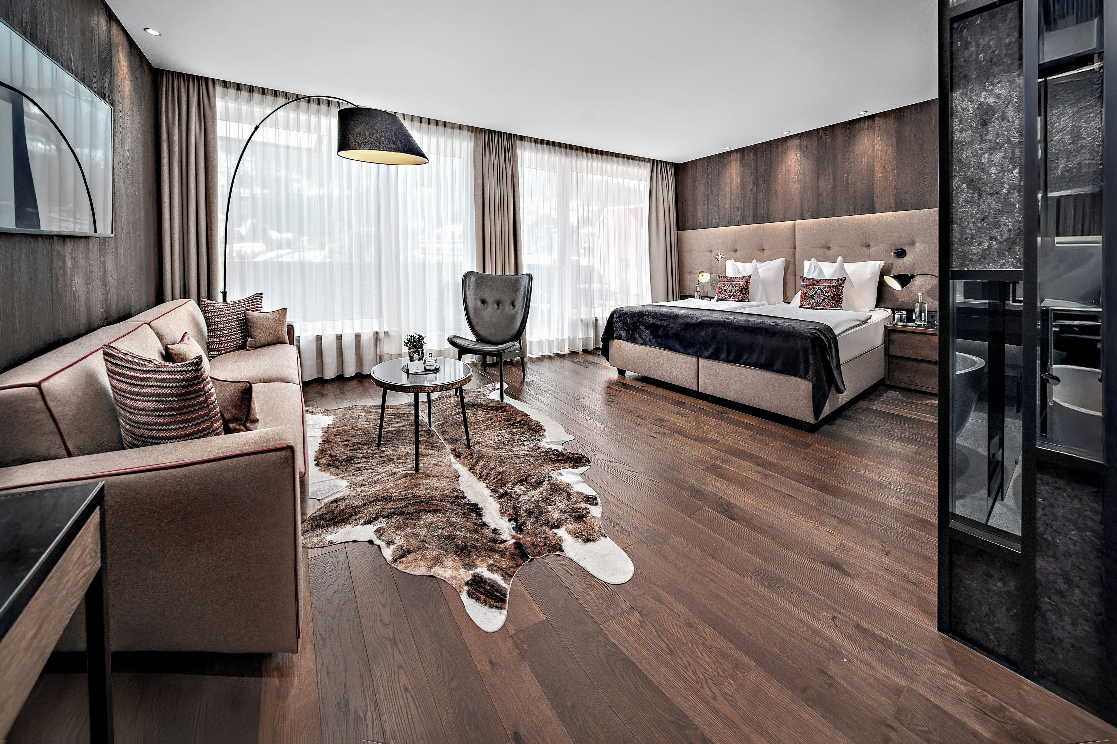 A modern hotel room with a sofa, a chair, a round coffee table, a bed with a headboard, and a large mirror.