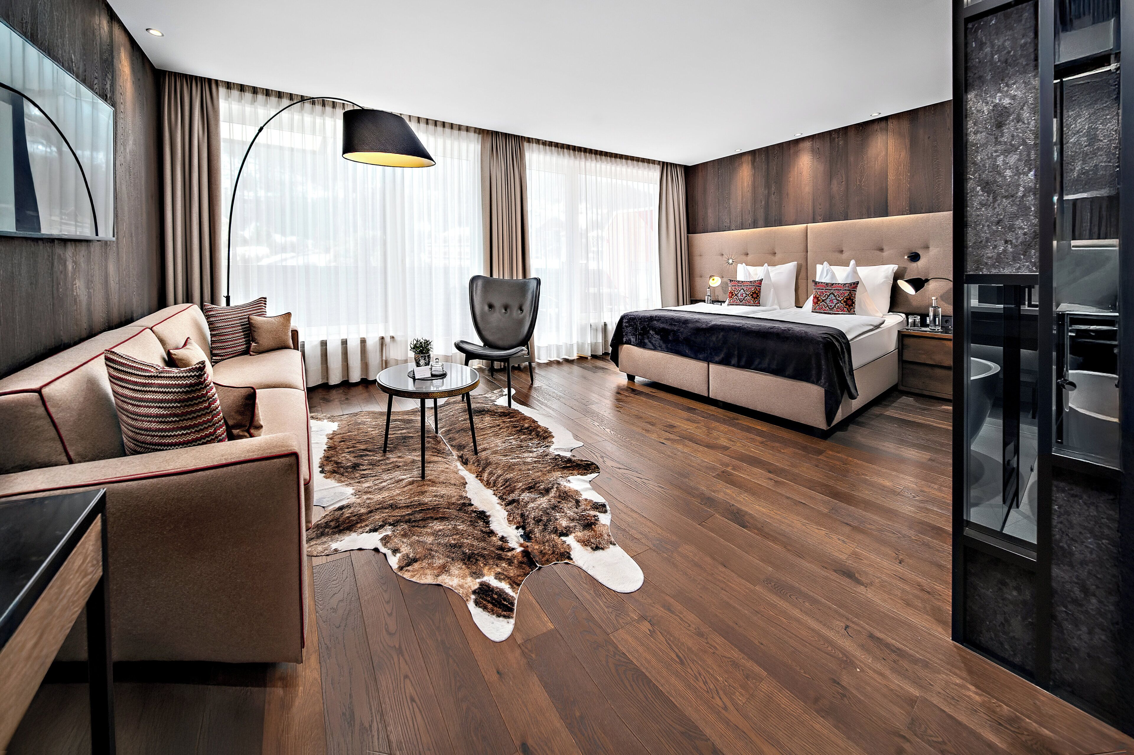 Une chambre d’hôtel moderne, équipée d’un canapé, d’un fauteuil, d’un lit, d’une petite table et d’une grande fenêtre avec des rideaux.