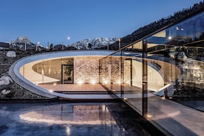 Een modern gebouw met een gebogen glazen gevel, een stenen binnenwand en een reflecterend waterkunstwerk ervoor.