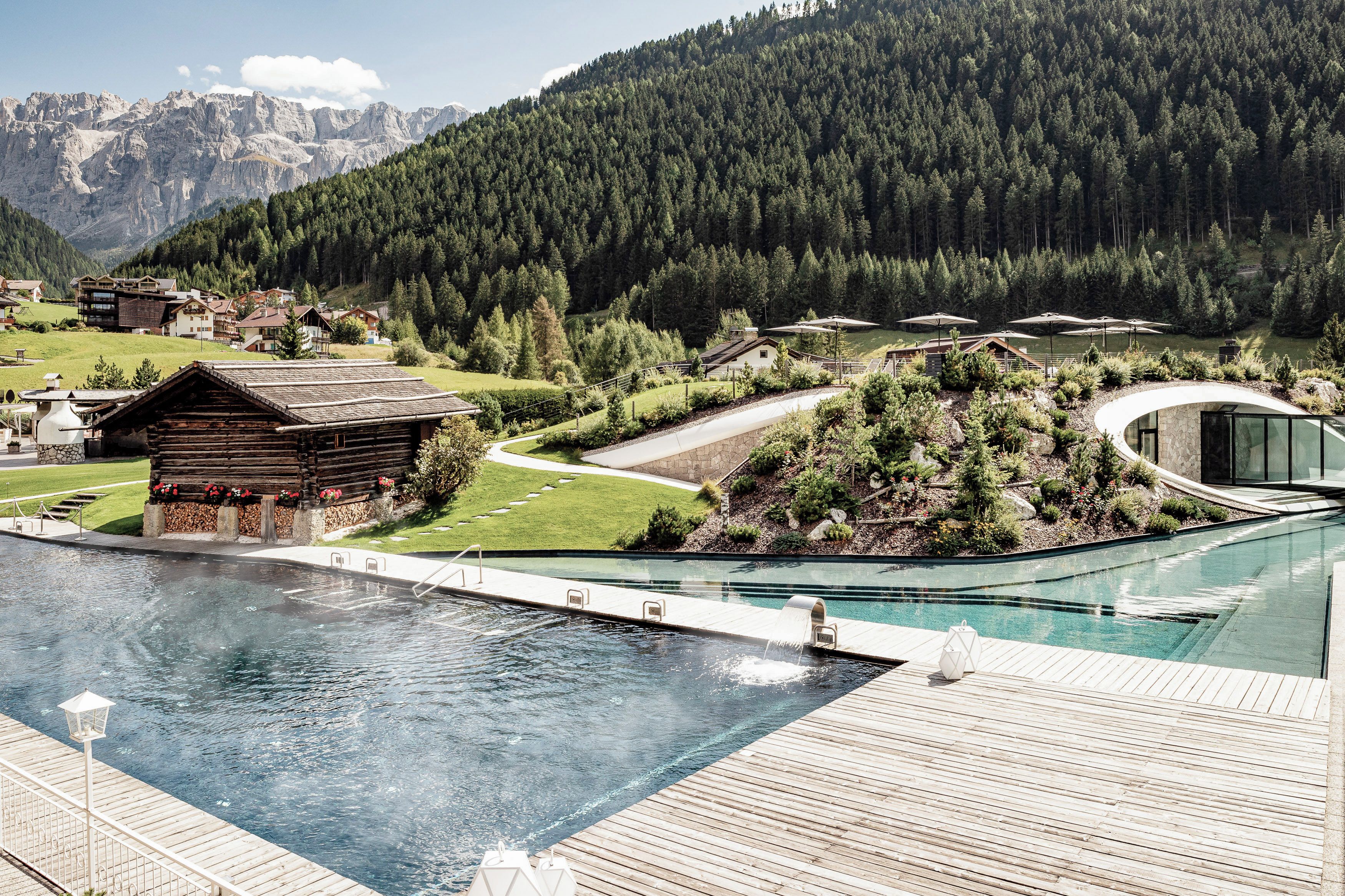 A pool area with a wooden deck, a small wooden cabin, and a modern building with a glass facade.