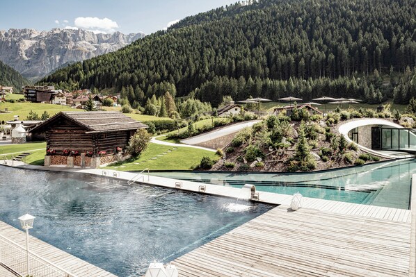 Un área de piscina con terraza de madera, una cabaña de madera pequeña y un edificio moderno con fachada de vidrio.