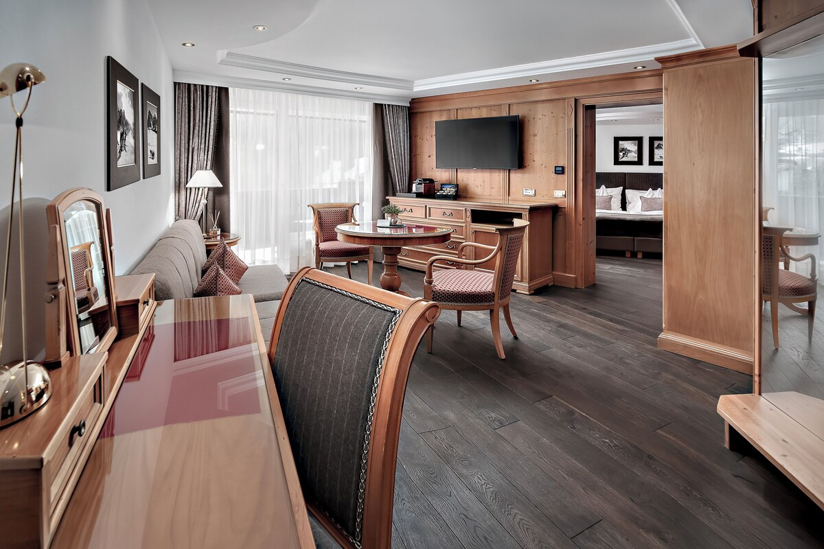 A hotel room with a dining area, a TV mounted on the wall, and a large window with curtains.