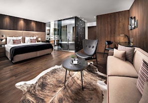 A modern hotel room with a bed, a sofa, a chair, a small table, and a glass partition.