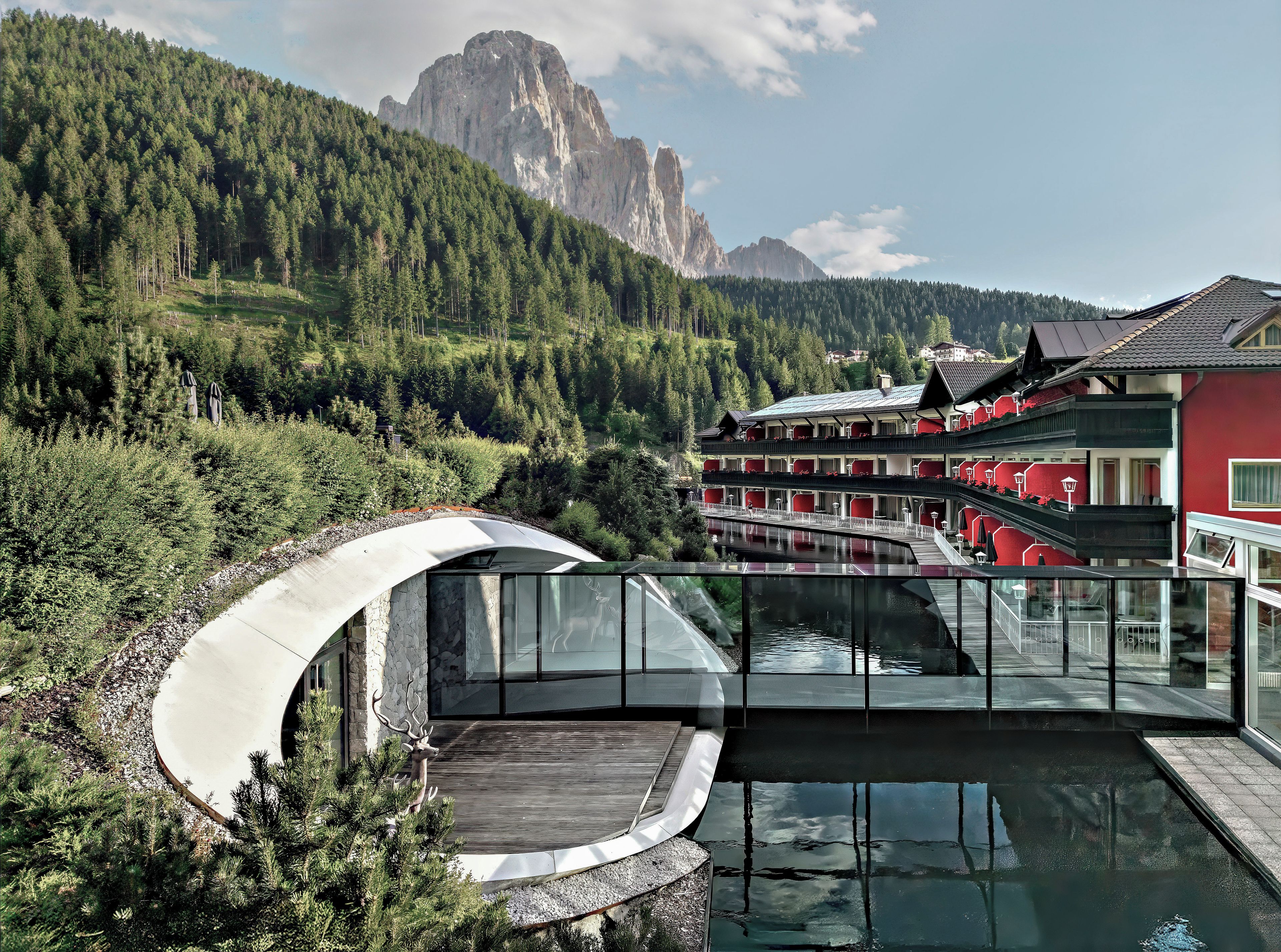 Un bâtiment moderne avec un pont de verre enjambant une étendue d’eau, entouré de collines boisées et avec une montagne en arrière-plan.