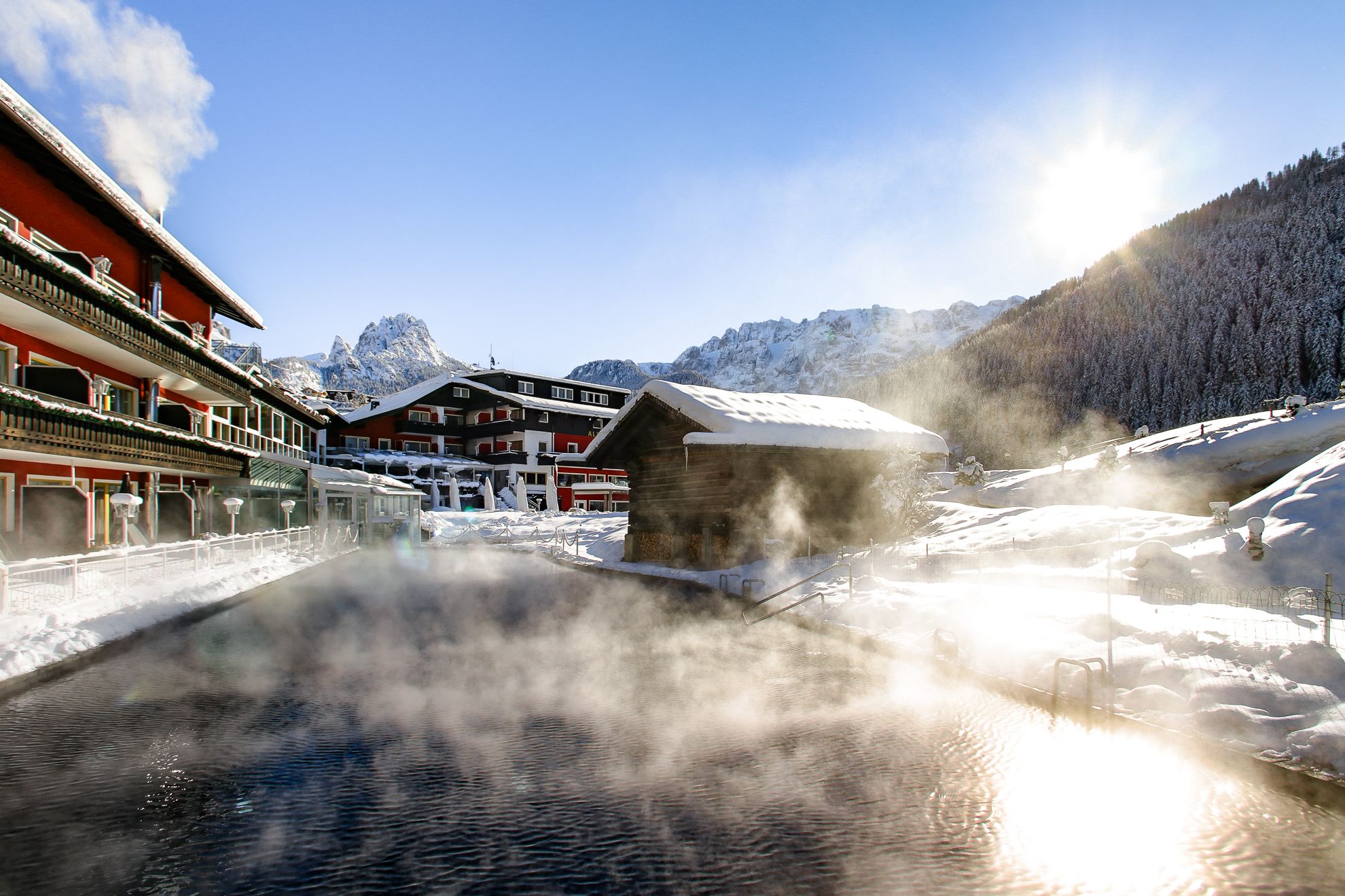 A traditional alpine hotel with a spa area, including a hot spring, surrounded by snow-covered mountains.