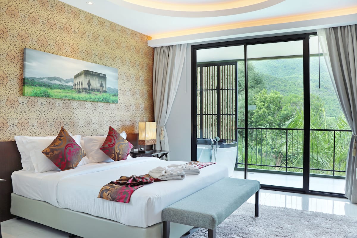 A modern hotel room with a large bed, a bench, a painting of a building, and a balcony with a view of greenery.