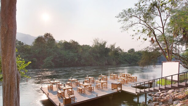 A wooden dock with tables and chairs extending over a calm river, surrounded by lush greenery and trees. - Mida Resort Kanchanaburi (Kanchanaburi)