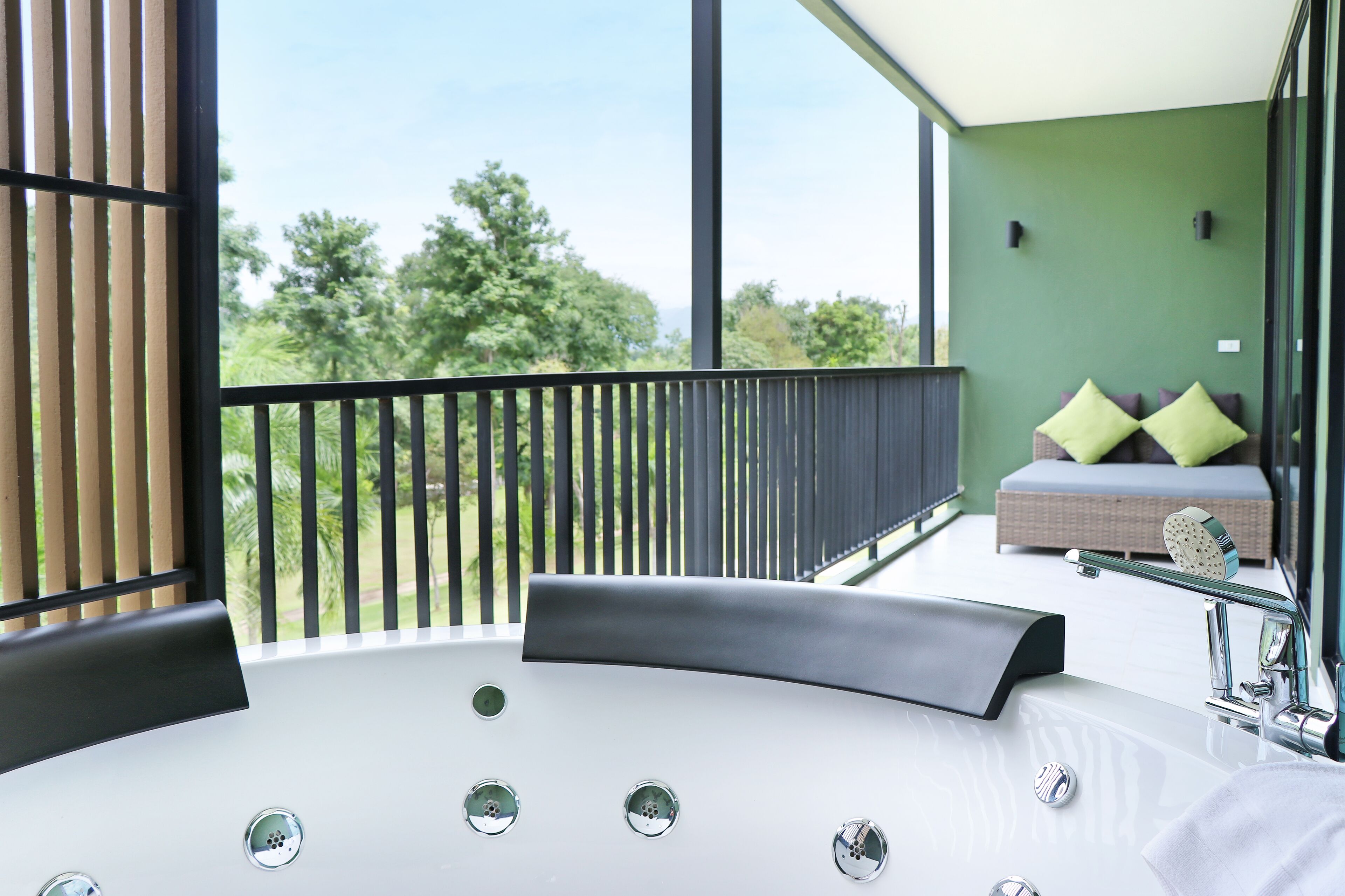 A modern bathroom with a large bathtub, a balcony with a view of trees, and a seating area with cushions.