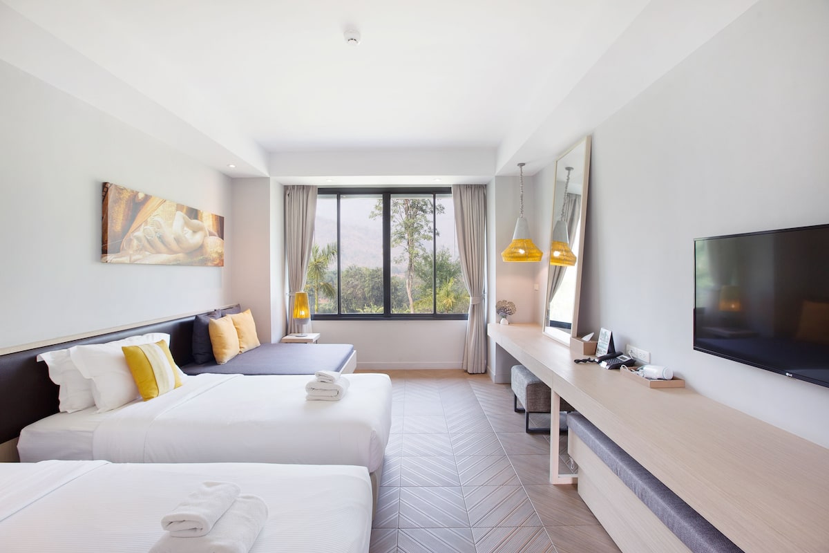 A hotel room with two beds, a TV, a desk, and a large window with a view of trees.