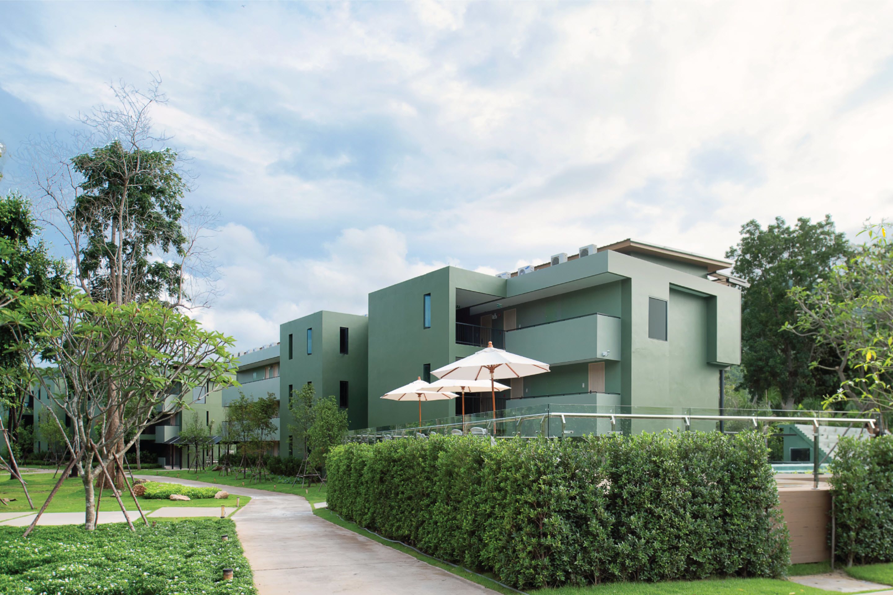 A modern building with a green facade, a pathway, and neatly trimmed hedges.