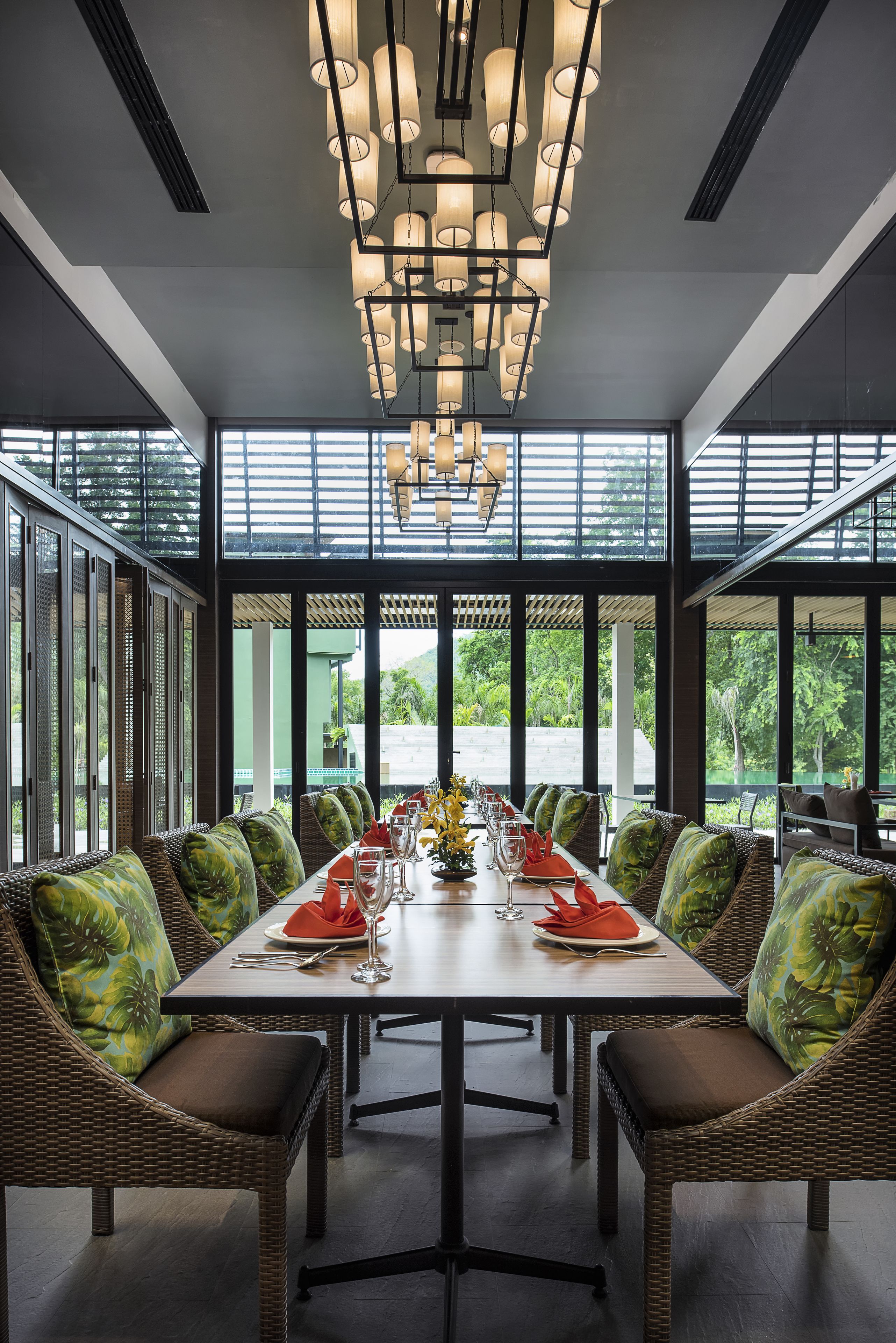 Un comedor con una mesa larga dispuesta para una comida, rodeado de sillas de mimbre con cojines verdes, y ventanas amplias que ofrecen vistas a la vegetación exterior.