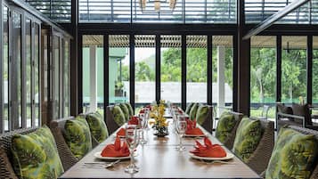Un comedor con una mesa larga dispuesta para una comida, rodeado de sillas de mimbre con cojines verdes, y ventanas amplias que ofrecen vistas a la vegetación exterior.