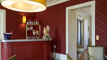 Un coin bar avec un mur rouge, une étagère à bouteilles encadrée de doré et un grand luminaire suspendu au plafond.