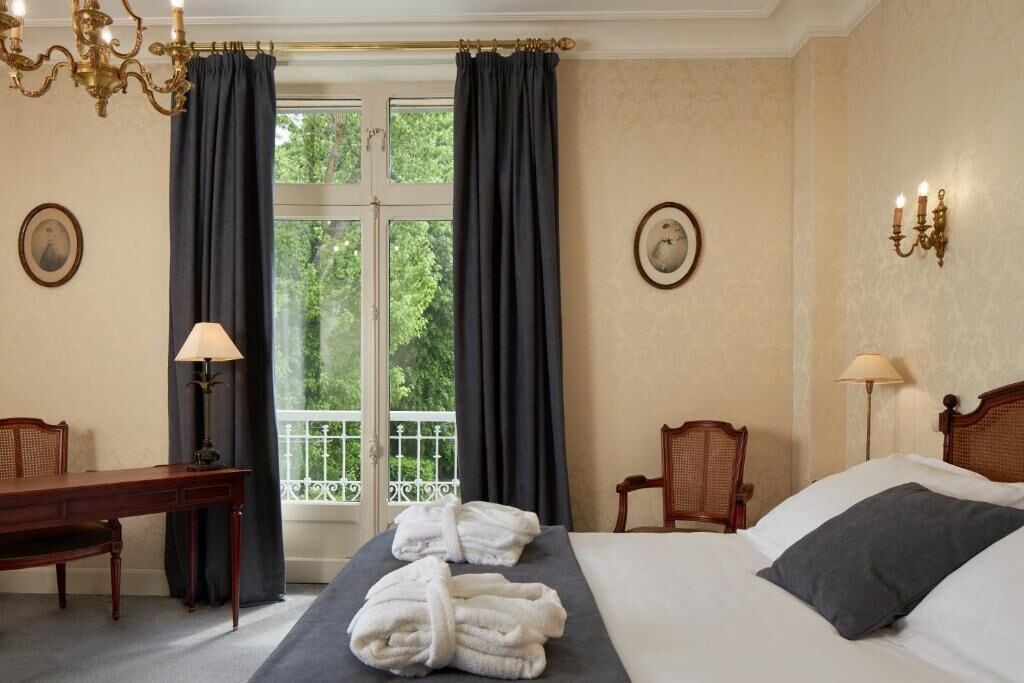 A hotel room with a bed, two chairs, a desk, a lamp, and a view of greenery through the window.