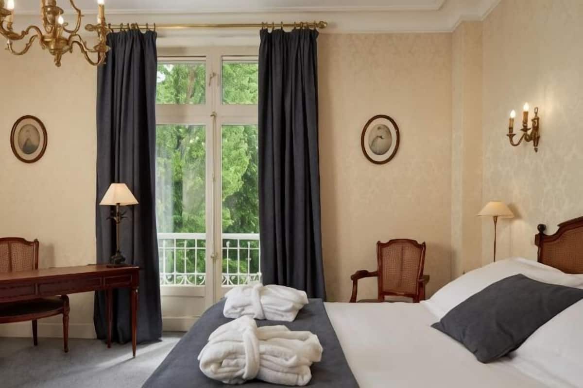Habitación de hotel con cama, dos sillones, escritorio, lámpara, candelabro y vistas por la ventana.