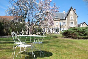 Un jardín con una mesa y sillas de metal blanco, un gran árbol en flor y un edificio con múltiples ventanas y techo a dos aguas.