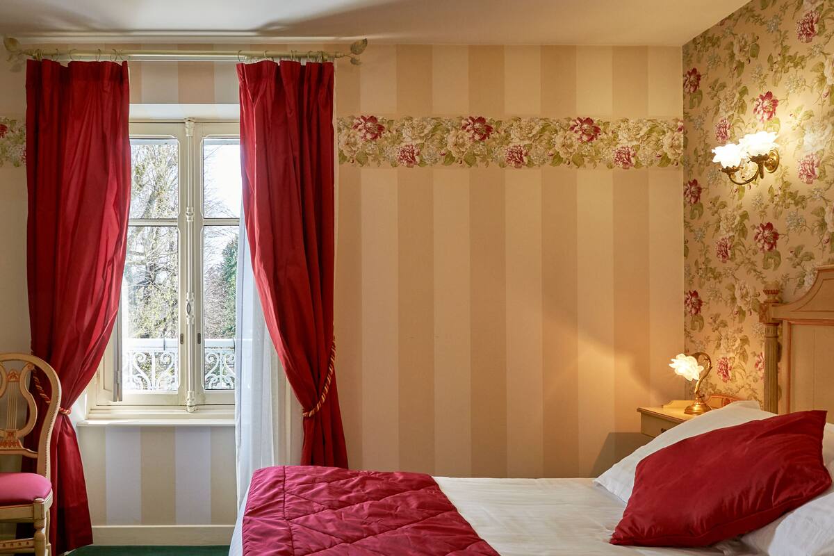 Un dormitorio con cama, cortinas rojas, una ventana, una mesita de noche y un papel tapiz floral.