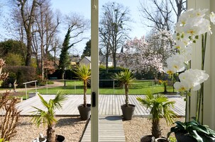 Una terraza de madera con plantas en macetas, vista a un jardín con árboles y césped, y una orquídea blanca en primer plano.