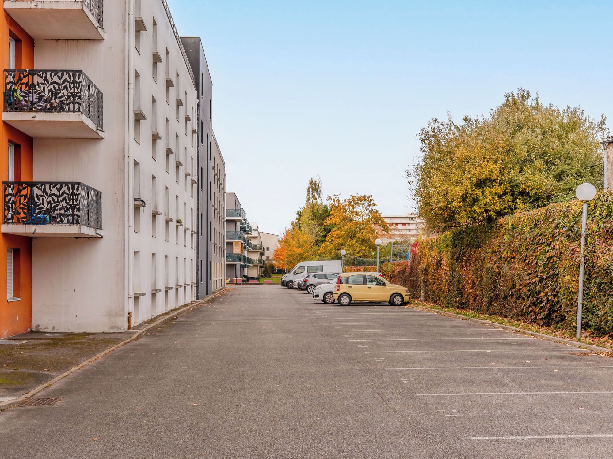 A parking lot with cars, a building with balconies, and autumn trees.