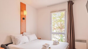 A hotel room with a bed, a nightstand, a lamp, a window with curtains, and a view of trees outside. - Sejours & Affaires Nantes La Beaujoire (Nantes)