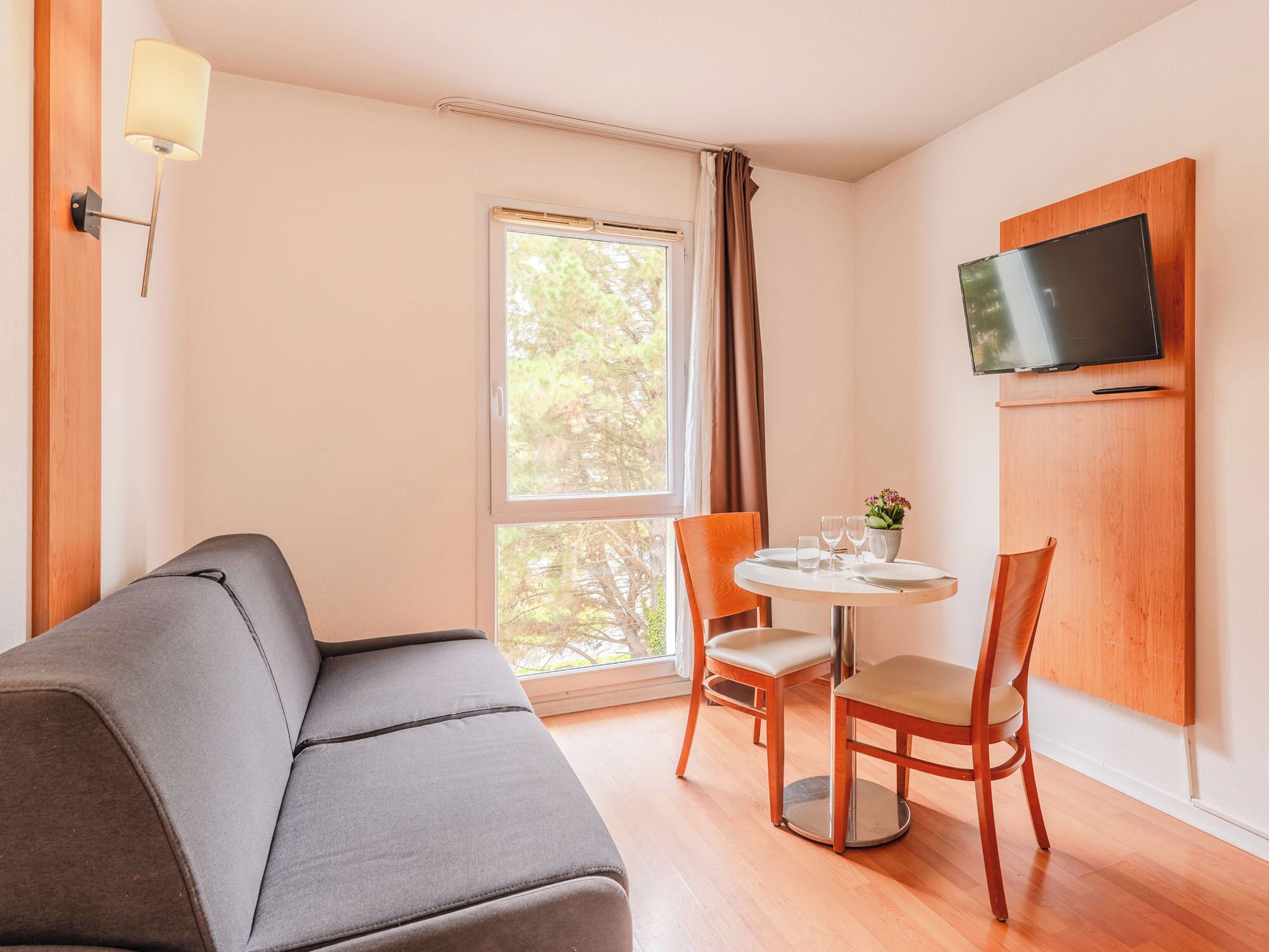 Una habitación moderna con un sofá gris, una mesa redonda de madera con dos sillas, un televisor montado en la pared y una ventana con cortinas.