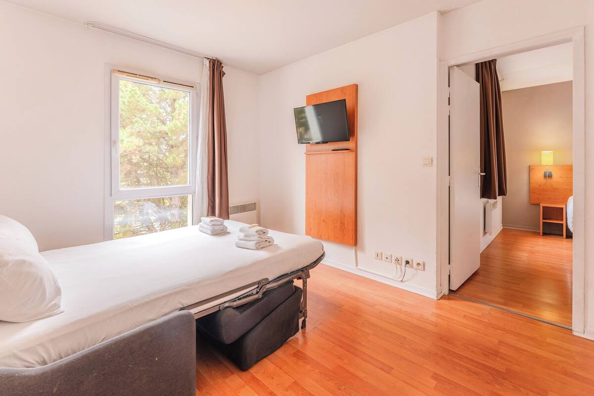 Ein kleines Hotelzimmer mit einem Einzelbett, einem an der Wand befestigten Fernseher und einem kleinen Tisch mit einer Lampe.