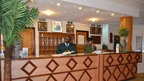 A reception desk with two staff members, a wooden panel design, framed pictures on the wall, and a potted plant. - Hotel Palm Beach (Ouagadougou)