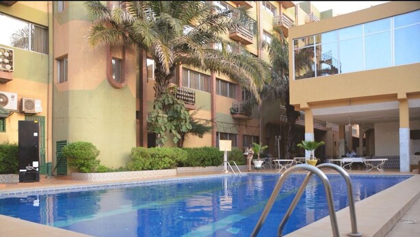 A swimming pool with a clear blue water, surrounded by a building with balconies and greenery. - Hotel Palm Beach (Ouagadougou)