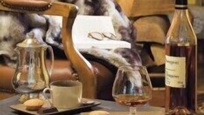 A cozy room with a fireplace, a leather armchair, a coffee table with a teapot, a glass, and a bottle of liquor.
