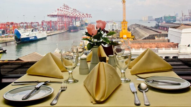 Mesa dispuesta para comer con vista a un puerto y grúas al fondo.