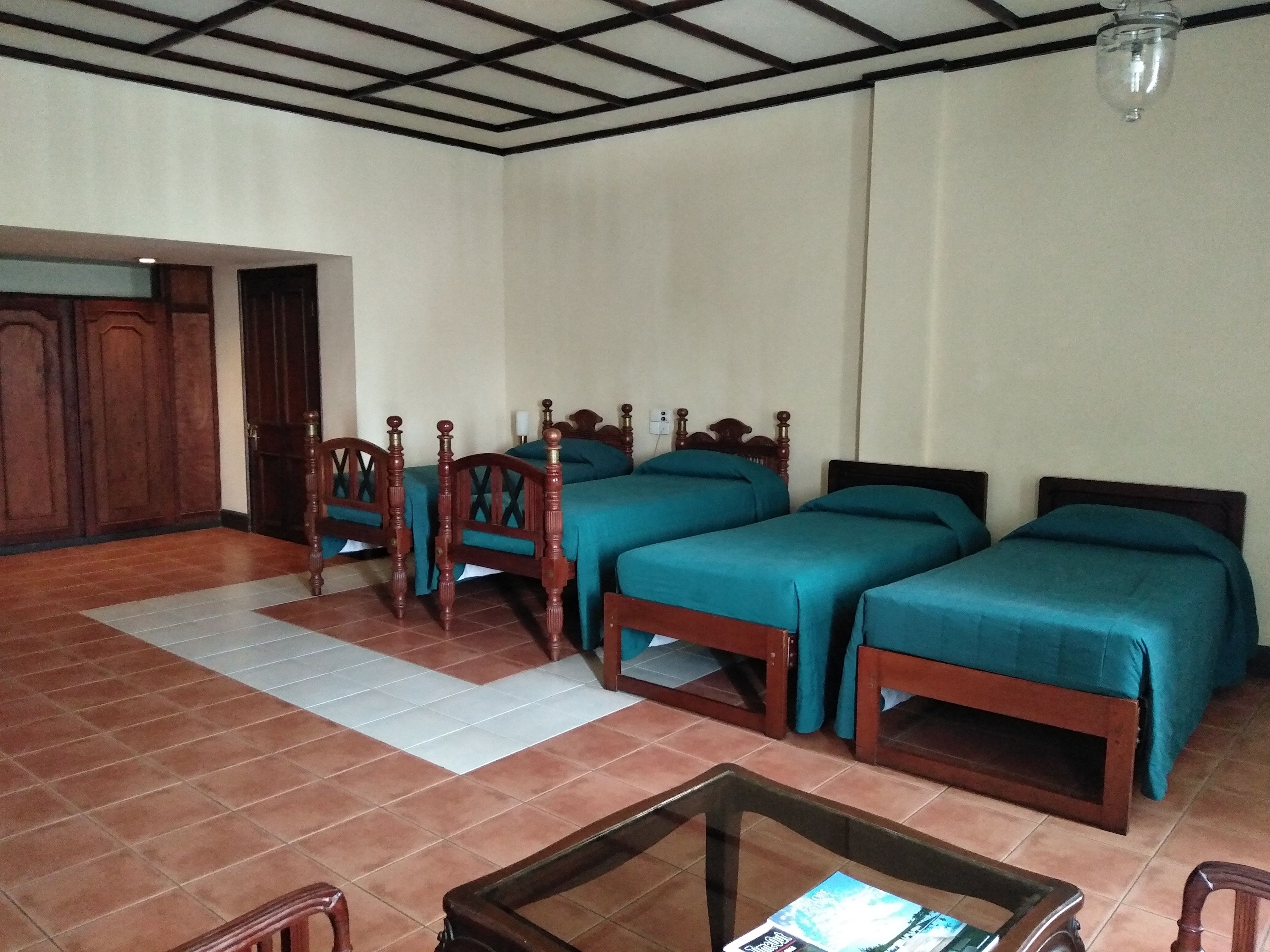 Habitación de hotel con cuatro camas de madera, suelo de baldosas, puerta de madera y techo con diseño de cuadrícula.