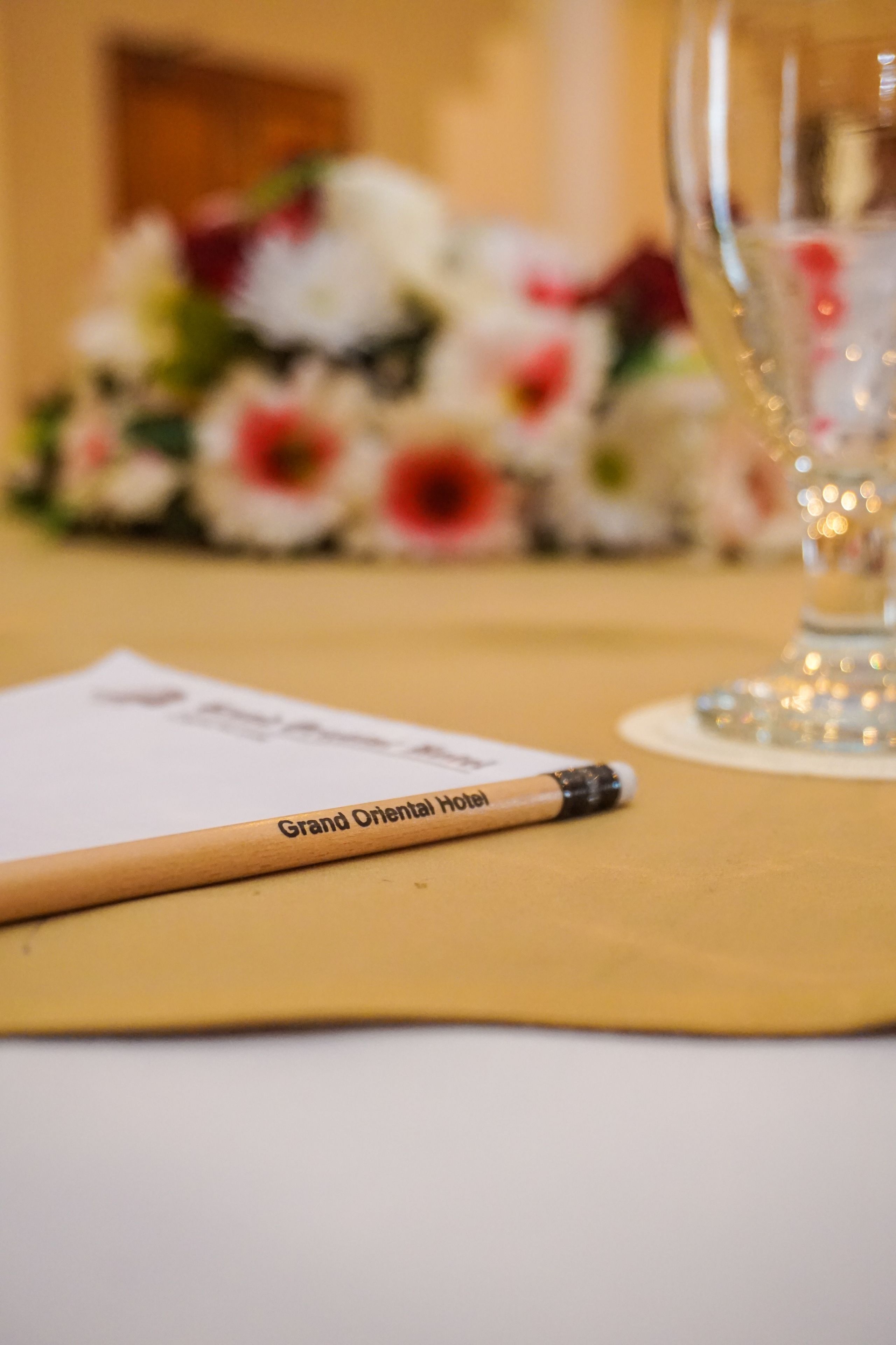 Une table avec un crayon provenant du Grand Oriental Hotel, un verre avec une bougie et un bouquet de fleurs en arrière-plan.