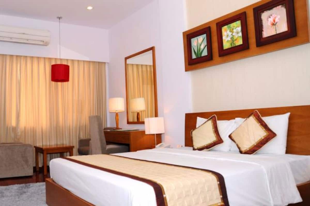 A hotel room with a bed, a desk, a chair, a lamp, a mirror, and a red hanging light fixture.