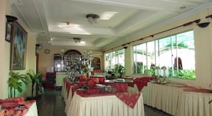 A restaurant with multiple tables covered in red tablecloths, a bar area with bottles, and large windows providing a view of greenery outside.