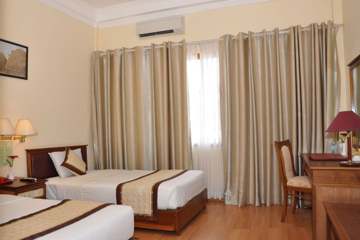 A hotel room with two beds, a desk, a chair, a lamp, and a window with curtains.