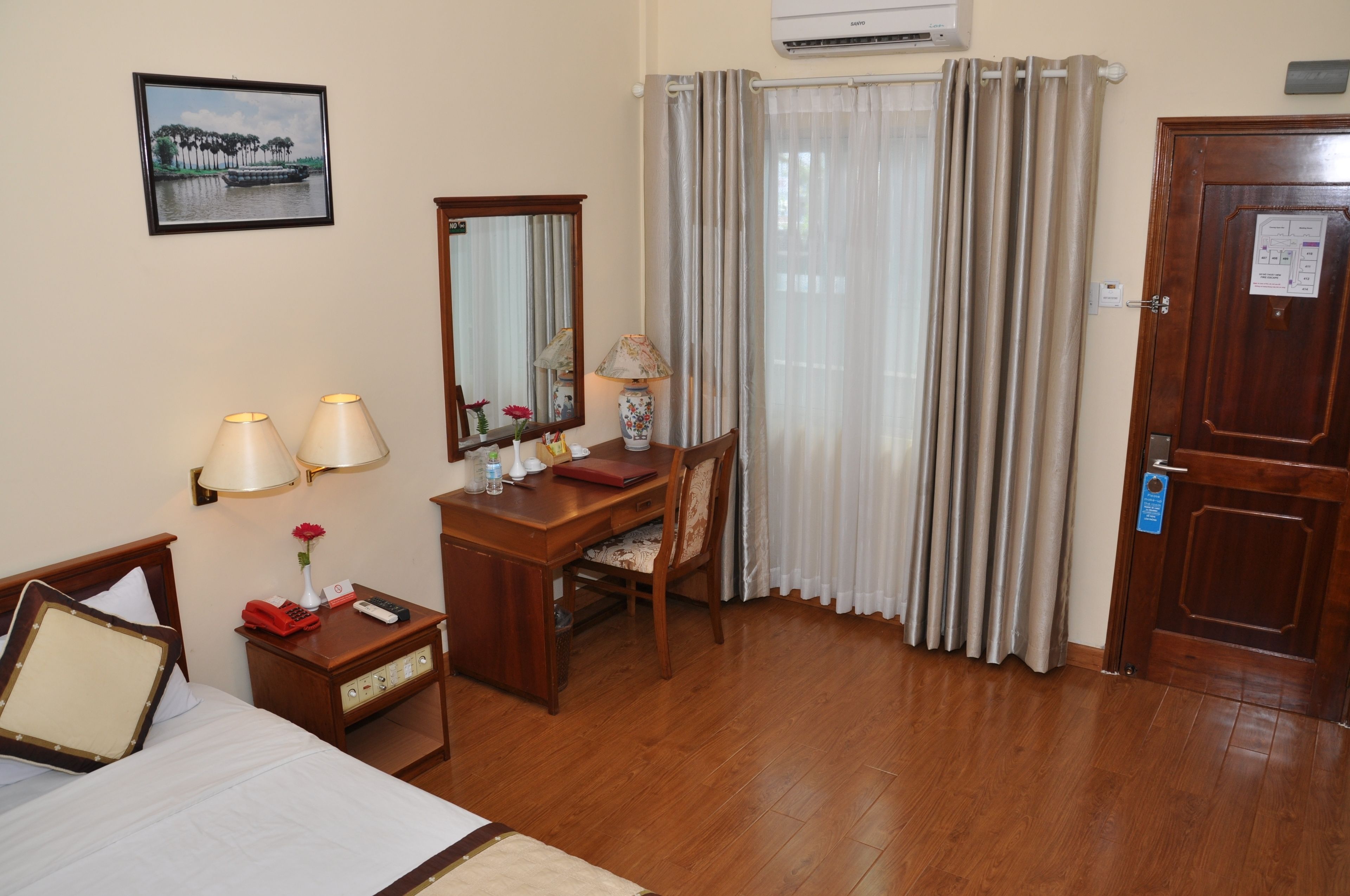 A hotel room with a bed, a desk with a chair, a lamp, a mirror, and a door.
