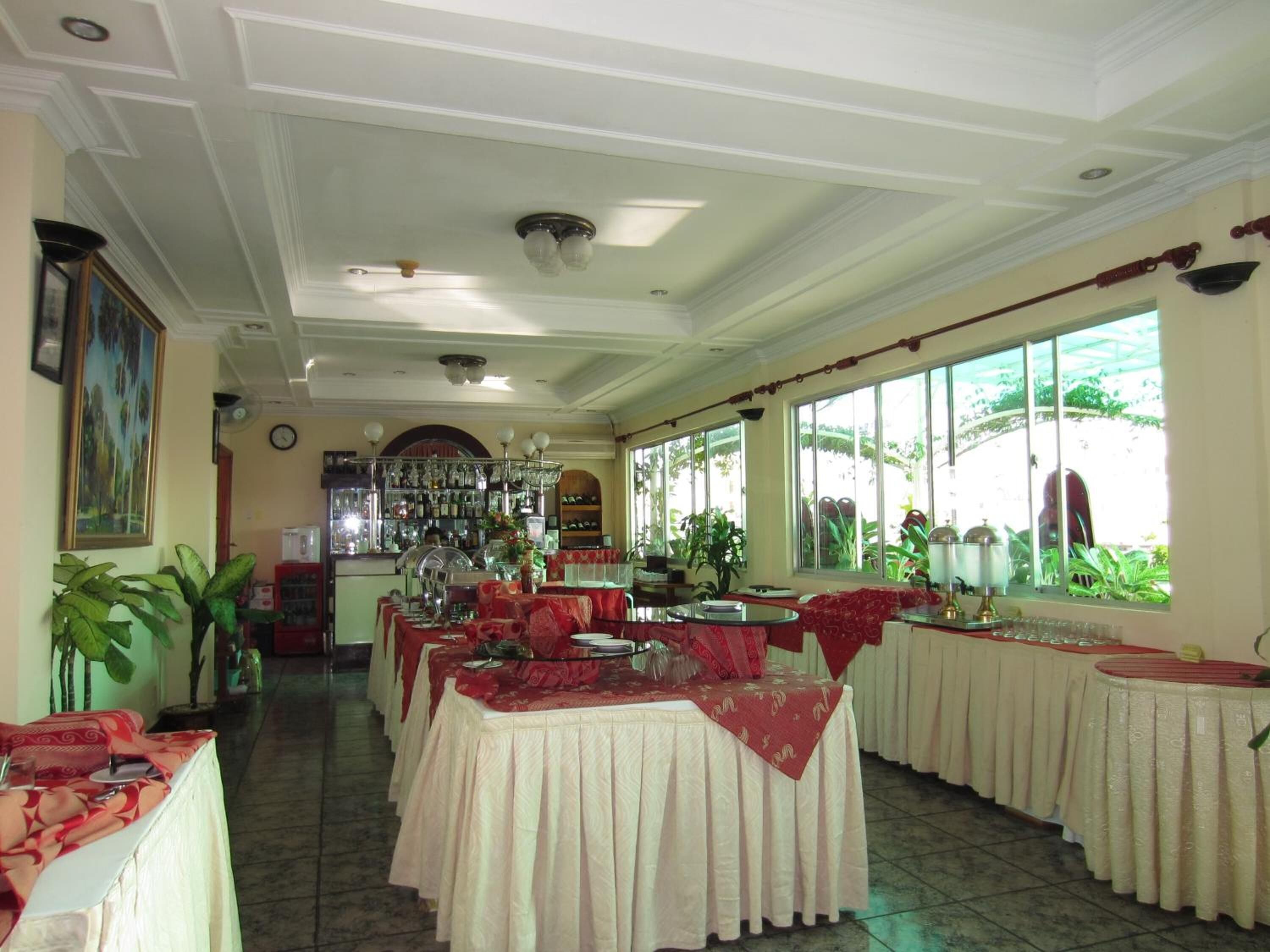 A restaurant with multiple tables covered in red tablecloths, a bar area with shelves of bottles, and large windows with a view of greenery.